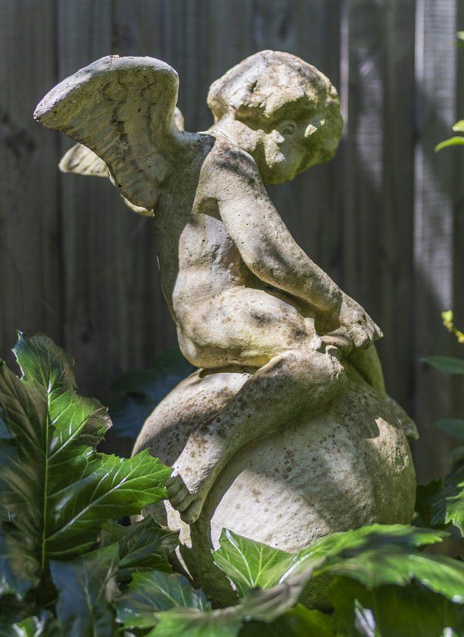 A cast cement garden sculpture of a winged putto, on a plinth